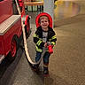 boots, boy, carpet, child, colorful_background, fire_hose, firefighter_costume, floor, fun, happy, indoor, jacket, kid, person, play_area, pretend_play, red_helmet, smiling, toy_fire_truck, young
