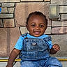 toddler, child, baby, smiling, denim_overalls, blue_shirt, stone_wall, concrete, barefoot, cute, happy, portrait, outdoor, sitting, young_child, person, infant, casual_clothing, expression, joyful