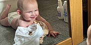 Theia is registered to the contest to win money with this photo: baby, infant, mirror, reflection, carpet, smiling, clothing, bedroom, wooden_furniture, bottle, lotion, diaper, floor, crawling, happy, indoor, child, person, baby_care, mirror_reflection