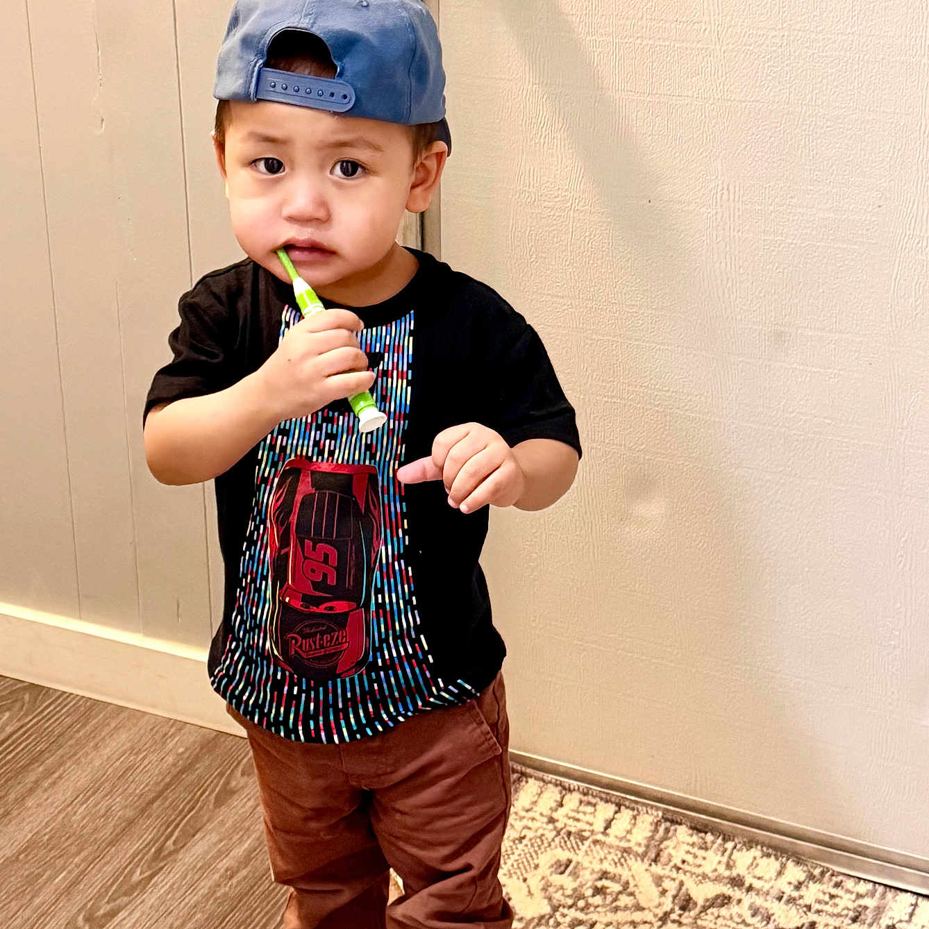 Thawng joined the competition — help win amazing prizes! toddler, child, barefoot, indoor, car_design, cap, blue_cap, black_tshirt, brown_pants, toothbrush, curious, standing, rug, door, wall, wood_floor, person, young_child, casual_clothing, home