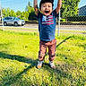 child, boy, grass, outdoor, happy, smiling, casual_clothing, cap, sneakers, daylight, fence, car, tree, shadow, cheerful, expression, standing, playful, nature, park