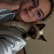 Blue a rejoint le concours — aidez-le/la à gagner de superbes lots ! person, cat, glasses, indoor, curly_hair, siamese_cat, hand, face, pet, affection, couch, sweatshirt, resting, closeup, portrait, animal, cozy, bond, relaxed, feline