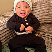 Ares is registered to the contest to win money with this photo: baby, child, infant, smiling, happy, portrait, sitting, indoor, couch, blanket, hat, knit_hat, black_clothing, onesie, pants, buttons, hands, face, home, cozy