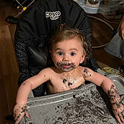 Marilyn is registered to the contest to win money with this photo: baby, child, high_chair, messy, chocolate, food, face_paint, arms, tray, indoor, wooden_floor, couch, toy, person, smudged, curly_hair, blue_eyes, expression, mess, cosco_kids