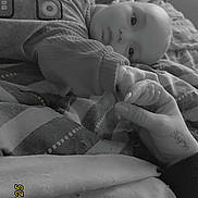 Nino a rejoint le concours — aidez-le/la à gagner de superbes lots ! baby, infant, hand, bed, blanket, pajamas, person, skin, face, holding_hands, resting, comfort, monochrome, indoor, cozy, sleepwear, blanket_pattern, human, calm, family