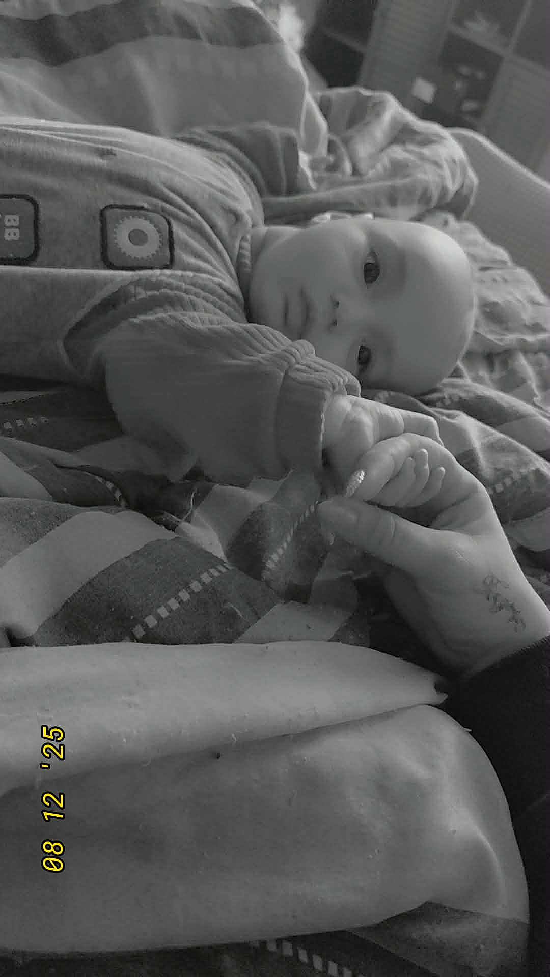 Nino a rejoint le concours — aidez-le/la à gagner de superbes lots ! baby, infant, hand, bed, blanket, pajamas, person, skin, face, holding_hands, resting, comfort, monochrome, indoor, cozy, sleepwear, blanket_pattern, human, calm, family