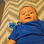 baby, blue_shirt, pacifier, blanket, smiling, lying_down, infant, child, bedroom, furniture, drawer, carpet, clip, happy, face, person, indoors, young_child, pattern, fabric