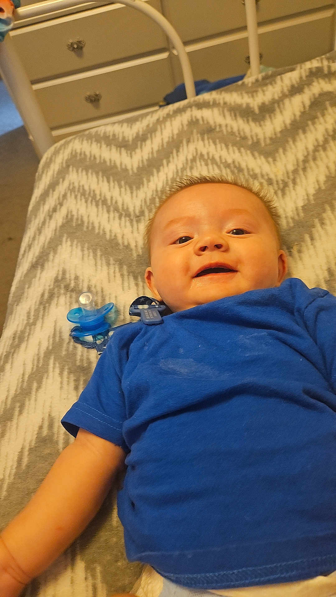 Austin Pierce joined the competition — help win amazing prizes! baby, blue_shirt, pacifier, blanket, smiling, lying_down, infant, child, bedroom, furniture, drawer, carpet, clip, happy, face, person, indoors, young_child, pattern, fabric
