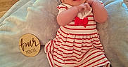 Nyla is registered to the contest to win money with this photo: baby, infant, four_months, smiling, happy, portrait, headband, red_dress, striped_clothing, bow, blanket, lying_down, indoor, wooden_floor, milestone, four_months_sign, face, hands, feet, family_photography