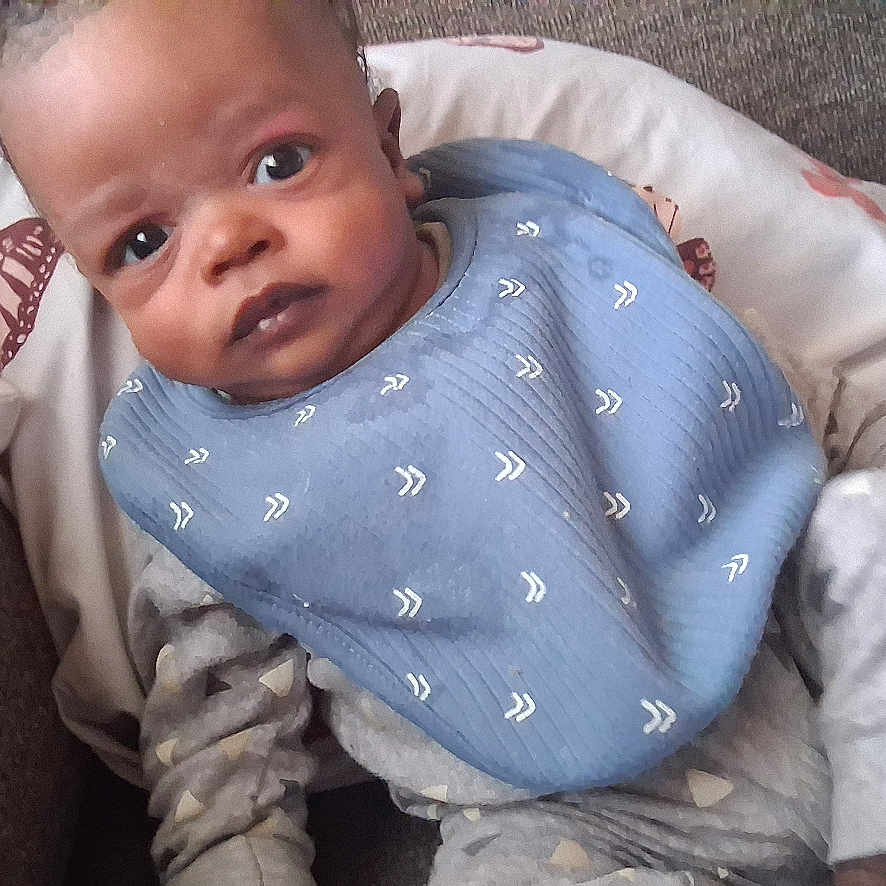 Alijah is registered to the contest to win money with this photo: baby, infant, child, portrait, face, wide_eyed, sitting, couch, sofa, cushion, bib, blue_clothing, bottle, baby_bottle, indoor, home, furniture, patterned_blanket, close_up, photography