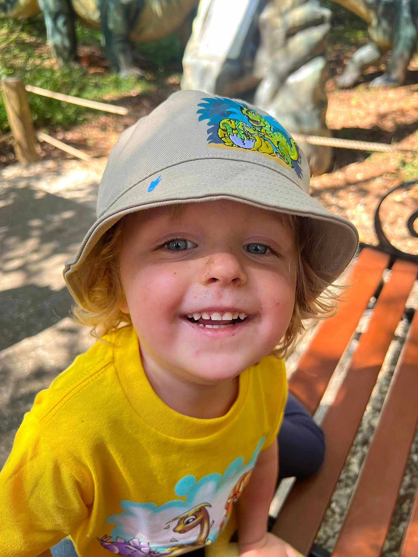 Axton joined the competition — help win amazing prizes! child, smiling, hat, bucket_hat, yellow_shirt, outdoor, bench, dirt, happy, portrait, daylight, cute, face, young, casual_clothing, nature, sunlight, blond_hair, playful, dinosaur_print