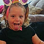 child, unicorn_headband, tongue_out, indoor, couch, pillow, black_shirt, playful, smile, person, hair, curly_hair, face, fun, headwear, living_room, happy, cute, closeup, seated