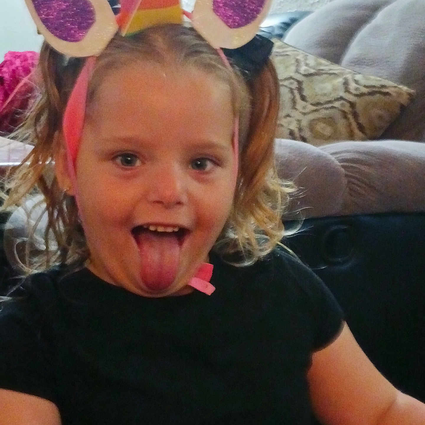 Evangelina is registered to the contest to win money with this photo: black_shirt, child, closeup, couch, curly_hair, cute, face, fun, hair, happy, headwear, indoor, living_room, person, pillow, playful, seated, smile, tongue_out, unicorn_headband