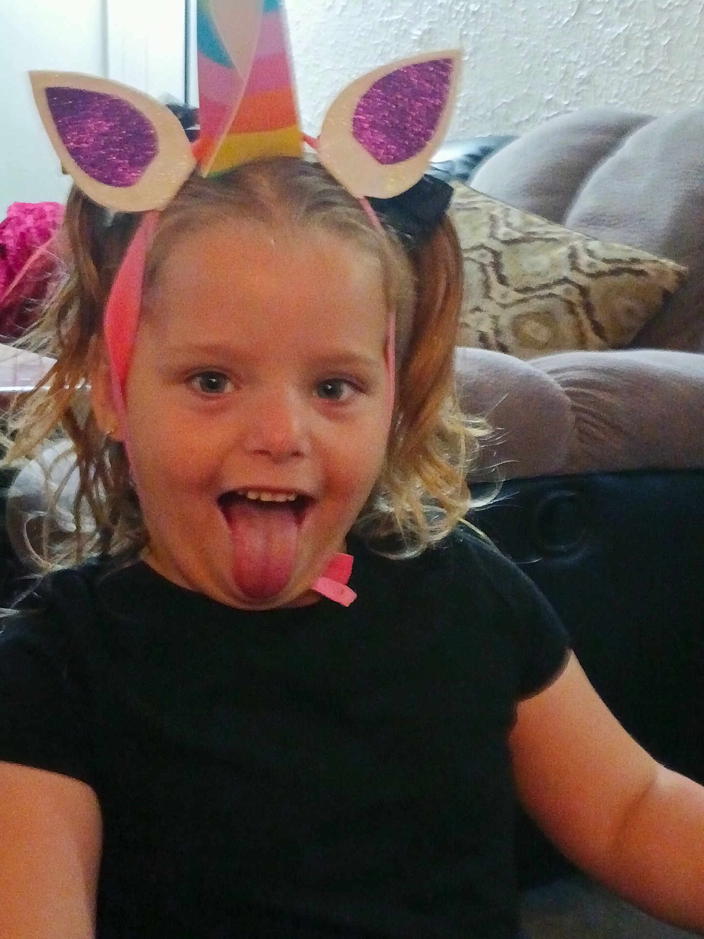 Evangelina is registered to the contest to win money with this photo: child, unicorn_headband, tongue_out, indoor, couch, pillow, black_shirt, playful, smile, person, hair, curly_hair, face, fun, headwear, living_room, happy, cute, closeup, seated