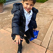 Hamzah Ali Saleh is registered to the contest to win money with this photo: child, boy, smiling, black_jacket, white_shirt, blue_bag, black_boots, outdoor, sidewalk, pavement, curb, greenery, building, window, playful, happy, walking, casual_clothing, daylight, person