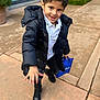 child, boy, smiling, black_jacket, white_shirt, blue_bag, black_boots, outdoor, sidewalk, pavement, curb, greenery, building, window, playful, happy, walking, casual_clothing, daylight, person
