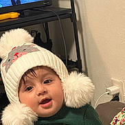 Faras Ali Saleh joined the competition — help win amazing prizes! baby, child, hat, pom_pom, white, green_sweater, indoor, face, cute, smiling, person, childhood, warm_clothing, home, furniture, television, cable, hand, portrait, cozy