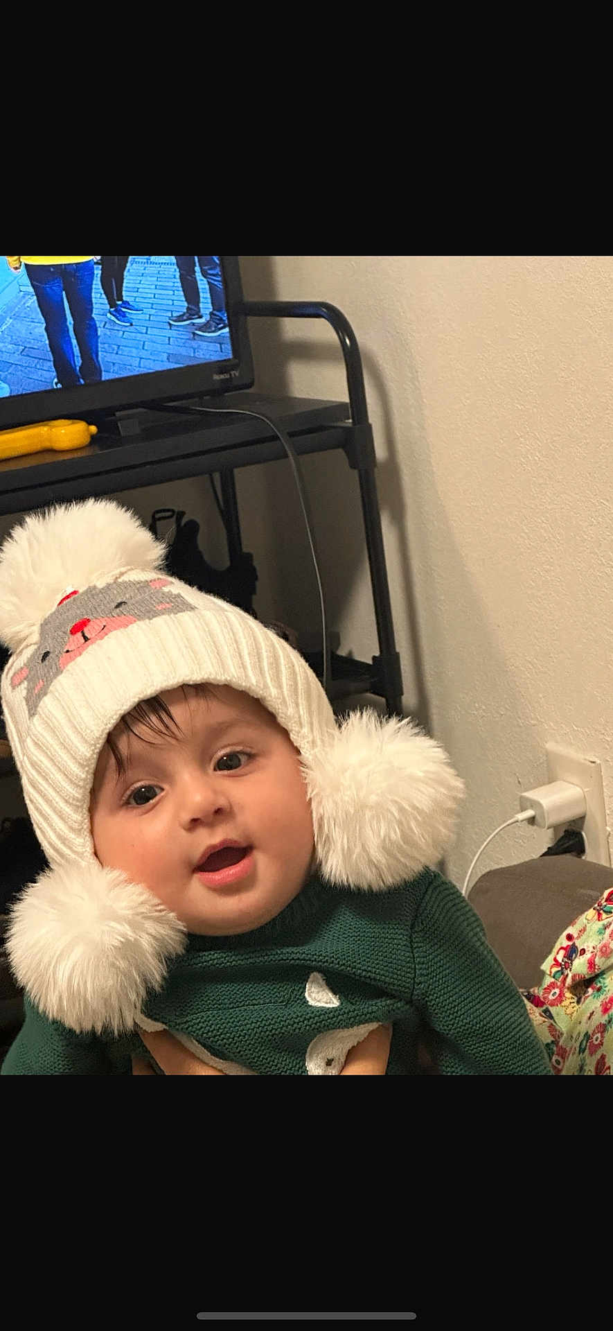 Faras Ali Saleh joined the competition — help win amazing prizes! baby, child, hat, pom_pom, white, green_sweater, indoor, face, cute, smiling, person, childhood, warm_clothing, home, furniture, television, cable, hand, portrait, cozy