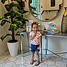 child, person, indoor, floor, tile_floor, mirror, plant, potted_plant, table, vase, flower_arrangement, casual_clothing, shorts, shirt, sandals, smiling, reflection, modern, decor, happy