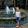 child, girl, fountain, water, outdoor, summer, casual_clothing, shorts, tshirt, sandals, paved_path, rocks, trees, urban, daylight, smile, playful, park, background, nature