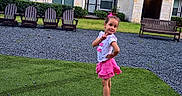 Linda is registered to the contest to win money with this photo: child, girl, posing, grass, lawn, pink_skirt, bow, apartment_building, residential, outdoor, daytime, smiling, happy, footwear, sandals, greenery, chairs, bench, sidewalk, casual_clothing