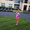 child, girl, posing, grass, lawn, pink_skirt, bow, apartment_building, residential, outdoor, daytime, smiling, happy, footwear, sandals, greenery, chairs, bench, sidewalk, casual_clothing
