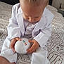 baby, bed, blond_hair, child, clothing, colorful, feet, hands, holding, home, indoor, looking_down, patterned_bedspread, person, sitting, soft_focus, sphere, toy, white_suit, young