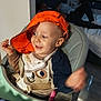 baby, bed, child, clothing, face, furniture, happy, headwear, high_chair, home, indoor, orange_hat, person, playful, relaxed, seat, smiling, table, toddler, toy