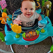 Liam is registered to the contest to win money with this photo: activity_jumper, baby, bright, carpet, child, clothing, colorful, cute, entertainment, face, fun, happy, indoor, infant, patterned_background, person, playful, sea_creatures, smiling, toys