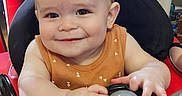 Hayes is registered to the contest to win money with this photo: baby, child, closeup, cute, face, fun, hands, happy, indoor, infant, orange_clothing, patterned_curtain, person, playtime, seat, smiling, steering_wheel, toy, walker, young_child