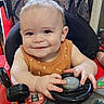 baby, child, closeup, cute, face, fun, hands, happy, indoor, infant, orange_clothing, patterned_curtain, person, playtime, seat, smiling, steering_wheel, toy, walker, young_child