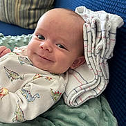 Hayes joined the competition — help win amazing prizes! baby, infant, smile, dinosaur_print, onesie, blanket, pillow, striped_cloth, face, head, comfort, couch, blue_couch, soft_texture, child, young, happy, portrait, indoor, closeup