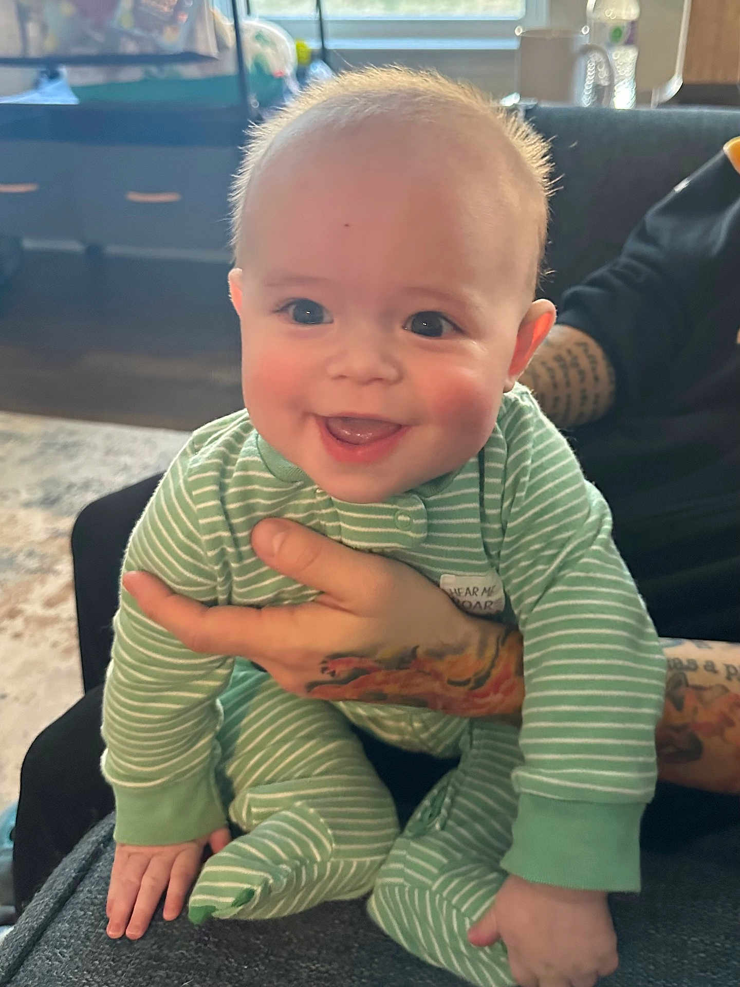 Hayes is registered to the contest to win money with this photo: baby, infant, smile, green_pajamas, tattoo, hand, indoor, furniture, couch, carpet, window, person, child, happy, face, cute, striped_clothing, sitting, young, cozy