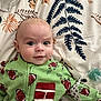 baby, child, infant, green_onesie, football_pattern, bedspread, floral_pattern, pacifier, face, eyes, smile, lying_down, blanket, fabric, indoors, cute, expression, clothing, person, portrait