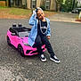 child, toy_car, pink, beaded_hair, necklace, jean_jacket, black_clothing, sneakers, outdoor, asphalt, suburban, house, trees, playful, smiling, portrait, daytime, casual, street, young_child
