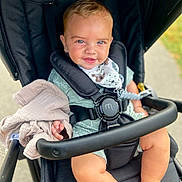 Cruz is registered to the contest to win money with this photo: baby, child, stroller, blue_eyes, blanket, smiling, outdoor, green_outfit, bib, black_stroller, cute, infant, person, seatbelt, happy, sitting, hand, foot, daylight, walk