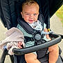 baby, child, stroller, blue_eyes, blanket, smiling, outdoor, green_outfit, bib, black_stroller, cute, infant, person, seatbelt, happy, sitting, hand, foot, daylight, walk