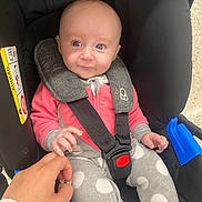 Maddison is registered to the contest to win money with this photo: baby, child, car_seat, hand, person, clothing, pink, gray, polka_dots, seatbelt, infant, cute, indoor, sitting, holding_hand, head, face, eyes, cozy, closeup