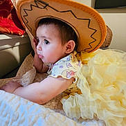 Amelia is registered to the contest to win money with this photo: baby, child, cowboy_hat, yellow_dress, bed, blanket, curious, indoor, portrait, soft_lighting, clothing, person, cute, toddler, home, decor, fabric, face, sleepwear, resting