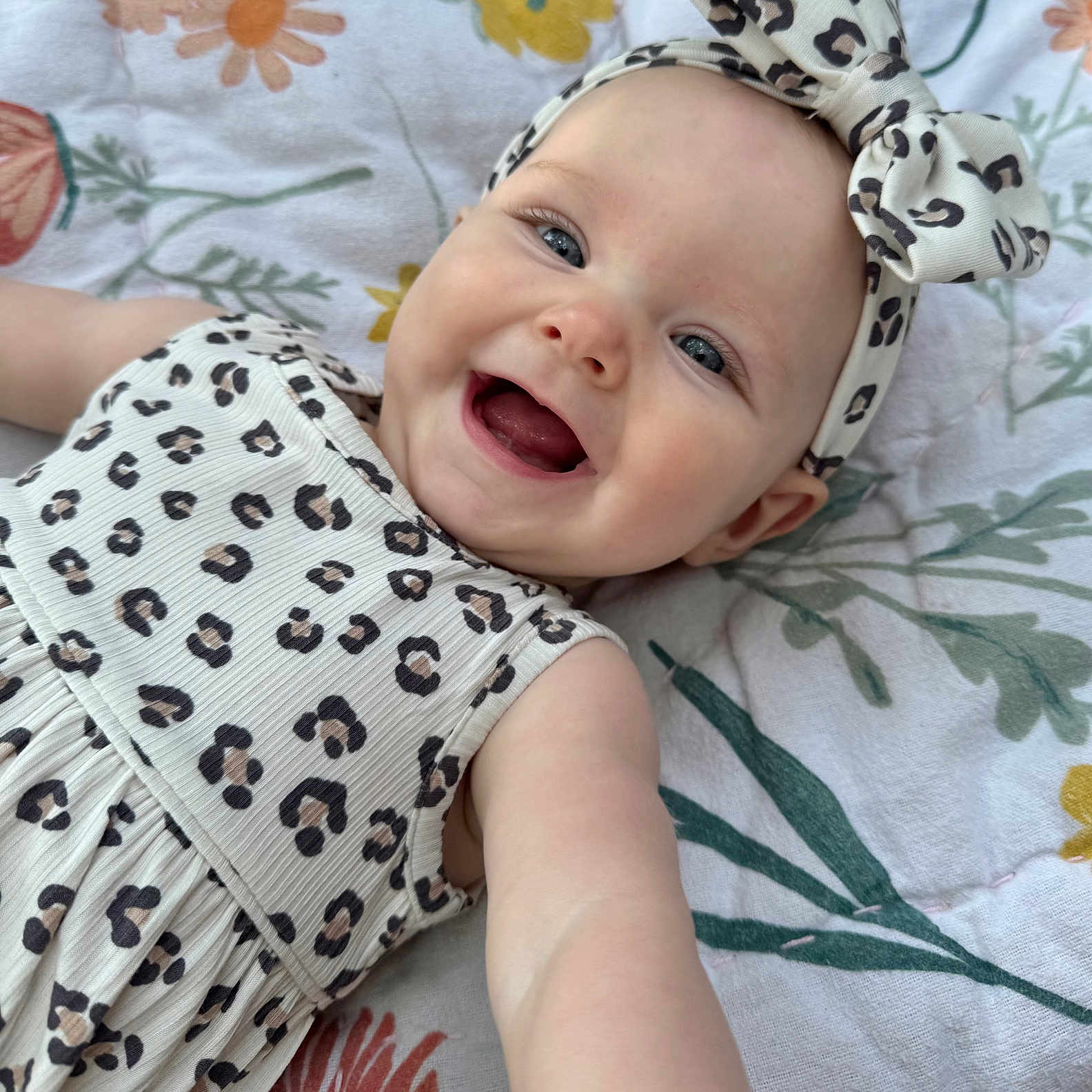 Riah is registered to the contest to win money with this photo: accessory, apparel, baby, blanket, blue_eyes, child, closeup, cute, dress, floral_pattern, happy, headband, infant, leopard_print, lying_down, person, portrait, reaching_out, skin, smiling