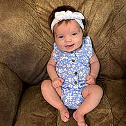 Wrenleigh is registered to the contest to win money with this photo: baby, infant, smiling, headband, blue_clothing, onesie, couch, brown_cushion, happy, sitting, cute, child, portrait, indoors, relaxed, skin, furniture, young_child, person, face