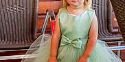 Lia participe au concours pour gagner de l'argent avec cette photo : child, toddler, dress, green_dress, handbag, white_handbag, sandals, bow, blonde_hair, blue_eyes, outdoor, wooden_wall, wicker_chair, floor, cute, fashion, portrait, person, standing, accessory