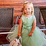 child, toddler, dress, green_dress, handbag, white_handbag, sandals, bow, blonde_hair, blue_eyes, outdoor, wooden_wall, wicker_chair, floor, cute, fashion, portrait, person, standing, accessory