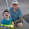 adult, bat, blue_clothing, cap, child, concrete, crocs, driveway, evening, family, glasses, glowing, outdoor, person, playful, shorts, smile, tattoo, toy, tshirt