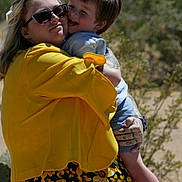Dean is registered to the contest to win money with this photo: affection, child, childrens_clothing, desert, family, floral_skirt, happy, hug, nature, outdoor, person, shorts, smile, sneakers, sunglasses, sunlight, tattoo, tree, woman, yellow_jacket