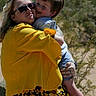 affection, child, childrens_clothing, desert, family, floral_skirt, happy, hug, nature, outdoor, person, shorts, smile, sneakers, sunglasses, sunlight, tattoo, tree, woman, yellow_jacket