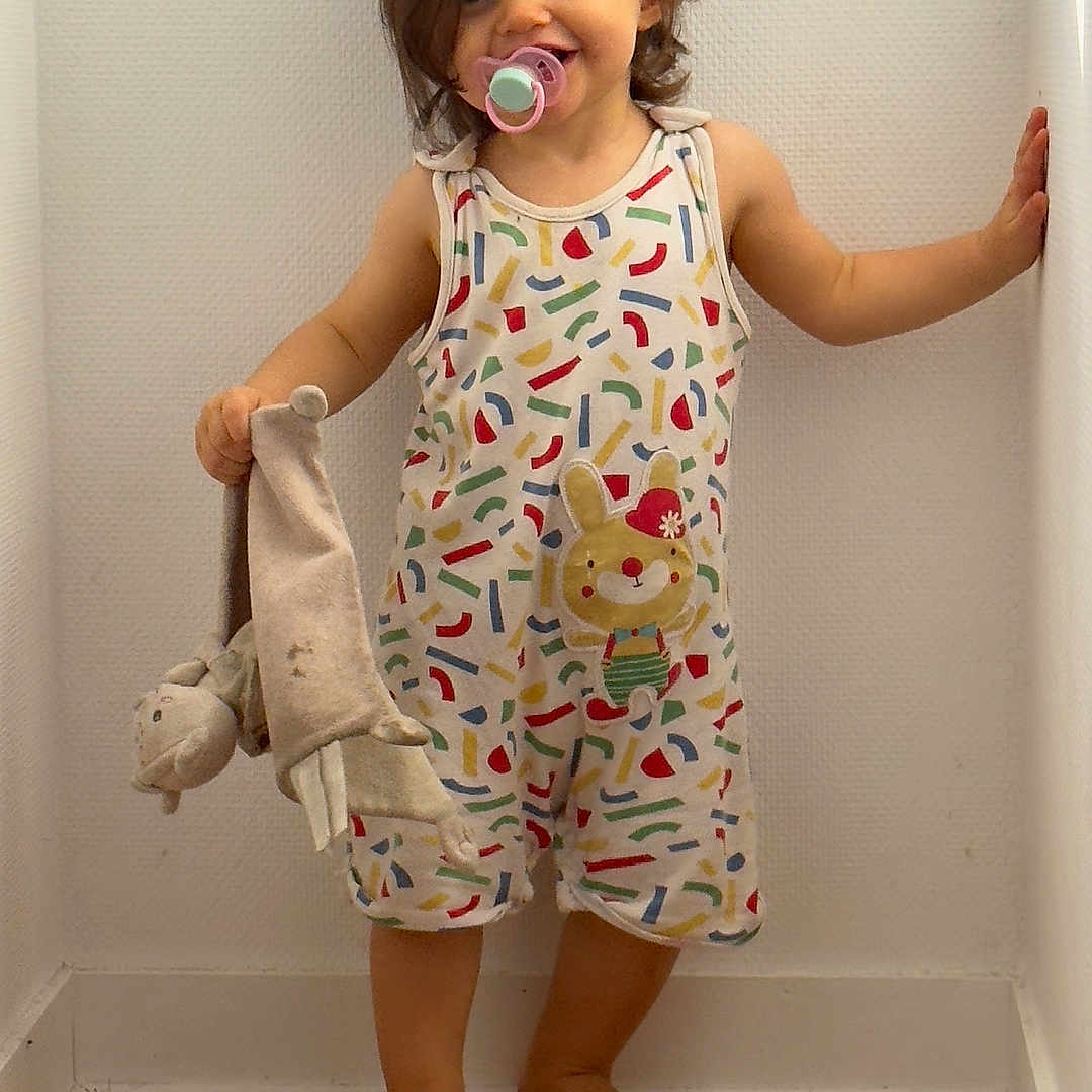 Layana a rejoint le concours — aidez-le/la à gagner de superbes lots ! baby, barefoot, child, clothing, corner, cute, happy, indoor, pacifier, person, playful, romper, short_hair, smiling, standing, stuffed_toy, toddler, toy, white_wall, wooden_floor