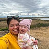 woman, baby, child, smile, happy, outdoor, nature, field, grass, cloudy_sky, lake, hat, knit_hat, yellow_sweater, holding, portrait, family, casual, daytime, person