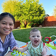 Jenna is registered to the contest to win money with this photo: woman, baby, smile, outdoor, grass, walker, toy, greenery, fence, sunlight, happy, child, person, nature, playtime, face, casual, daytime, family, portrait