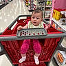 Jenna is registered to the contest to win money with this photo: baby, shopping_cart, store_aisle, child, pink_clothing, bib, socks, red_cart, indoor, retail, sports_equipment, curious, person, face, floor, shelf, shopping, baby_seat, grocery_store, expression